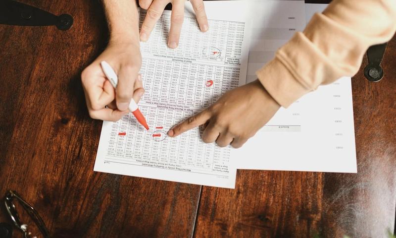 Two hands reviewing and marking financial documents with a red pen on a wooden table, top view.