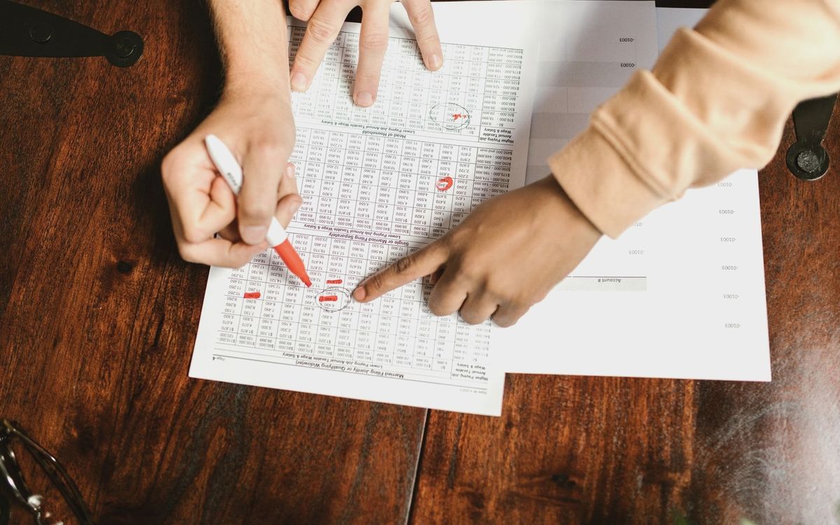 Two hands reviewing and marking financial documents with a red pen on a wooden table, top view.