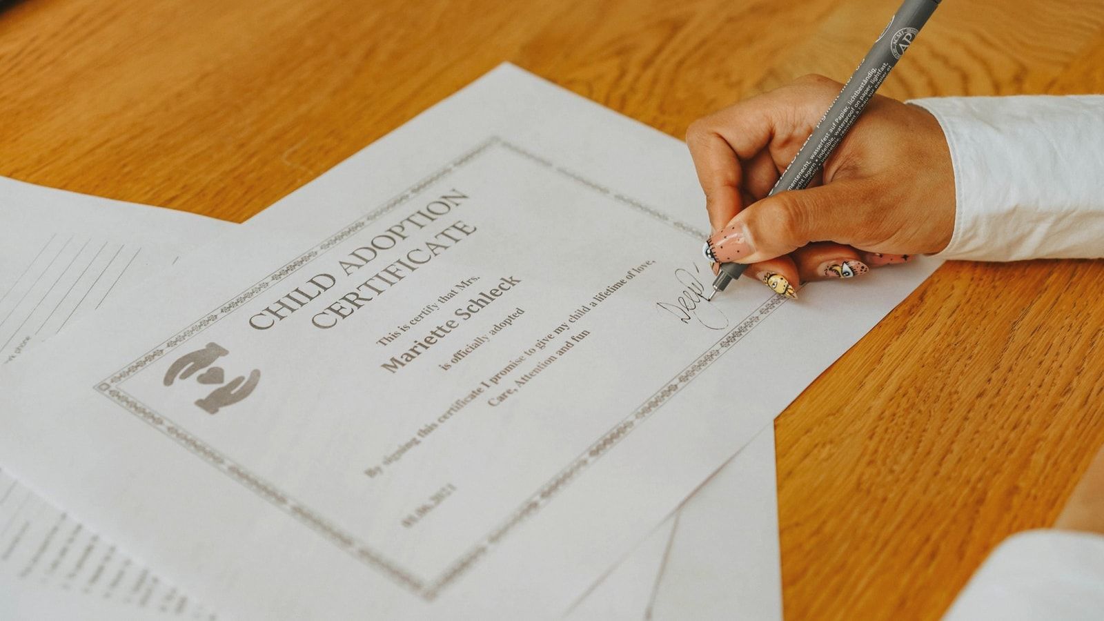 A close-up of a hand signing a child adoption certificate with a pen on a wooden table.