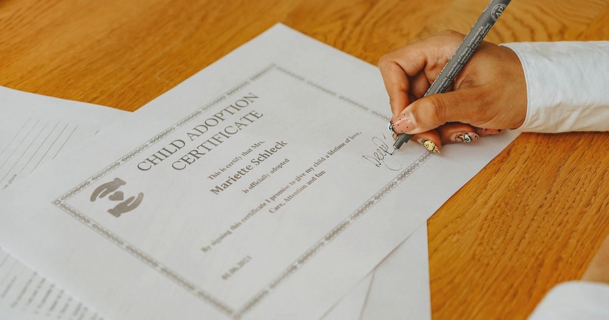 A close-up of a hand signing a child adoption certificate with a pen on a wooden table.