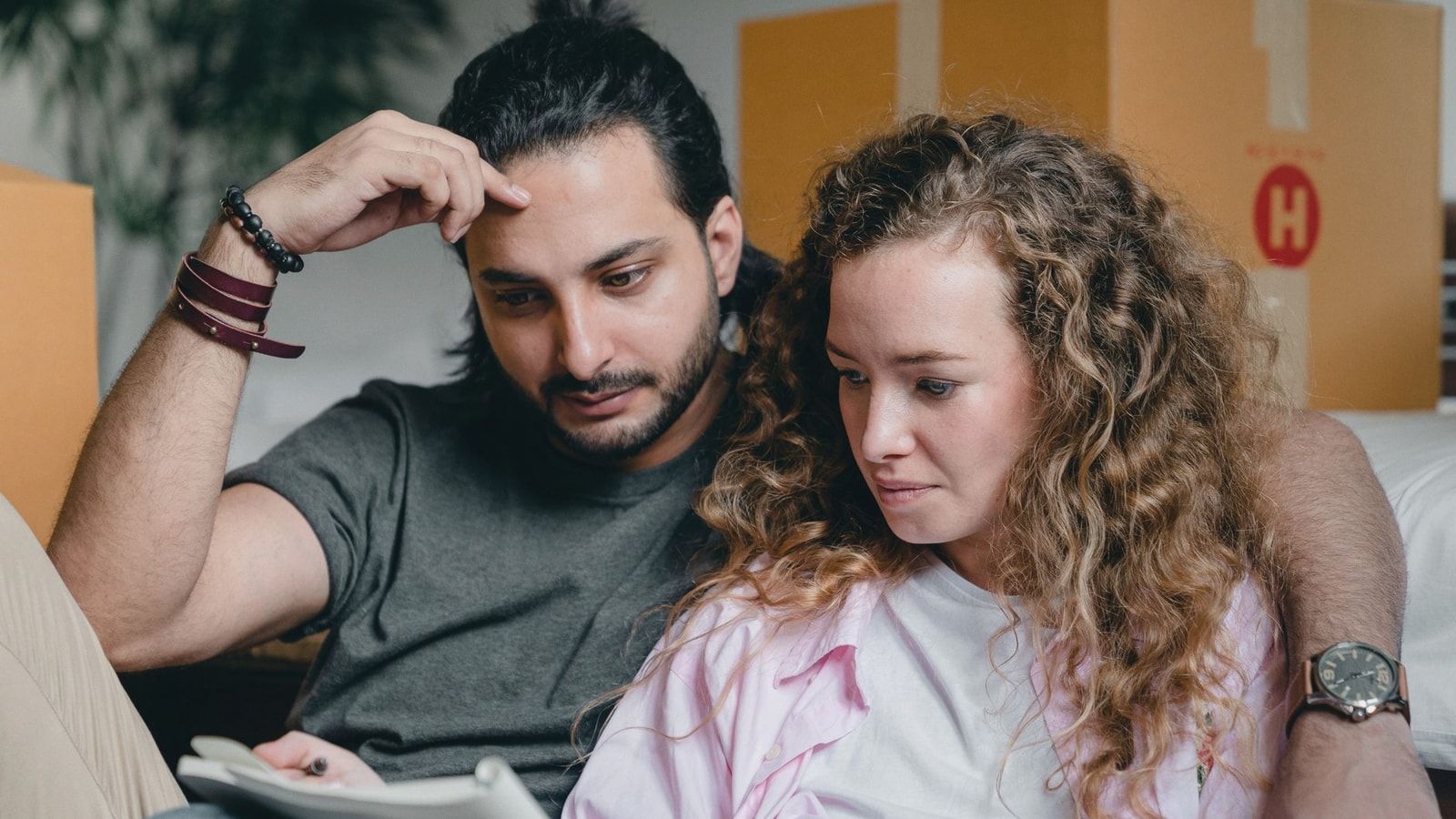 Focused male and female in casual wear sitting near boxes and looking through notebook thoughtfully while moving house