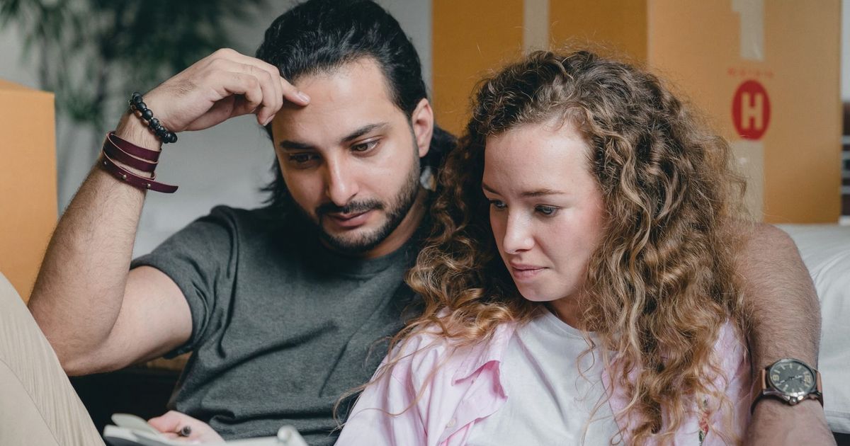 Focused male and female in casual wear sitting near boxes and looking through notebook thoughtfully while moving house
