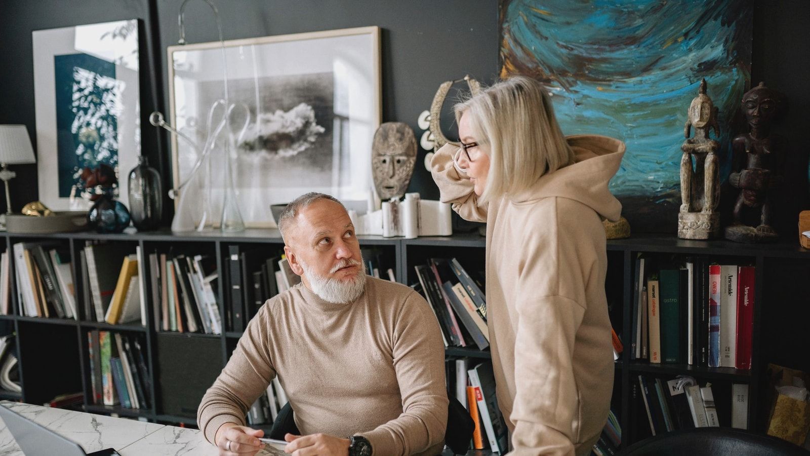 Elderly couple discussing something at a marble table with books and laptop, surrounded by art and sculptures.