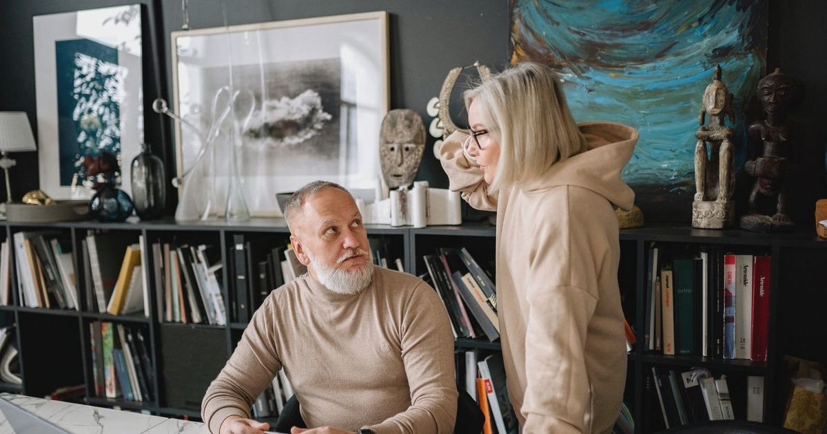 Elderly couple discussing something at a marble table with books and laptop, surrounded by art and sculptures.