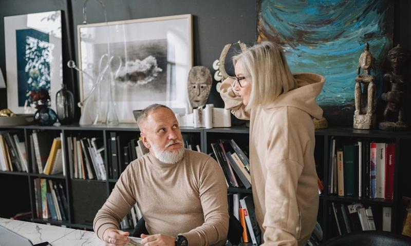 Elderly couple discussing something at a marble table with books and laptop, surrounded by art and sculptures.