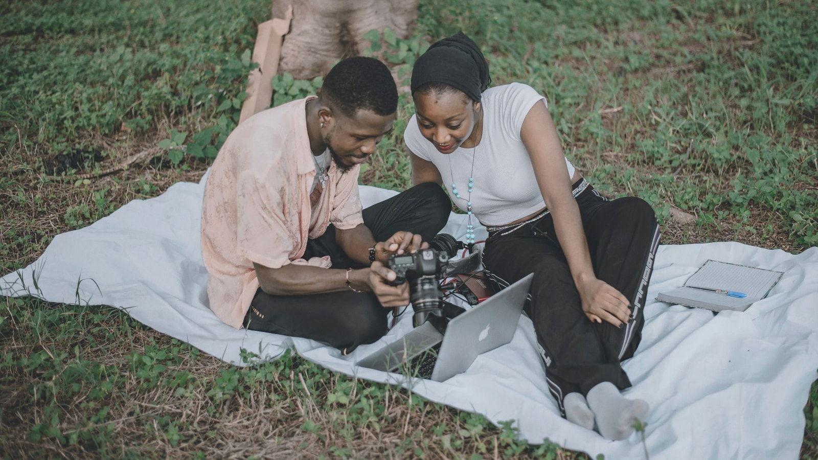 A couple sitting on a picnic blanket in a grassy area reviewing photos on a camera and laptop, enjoying leisure time outdoors.