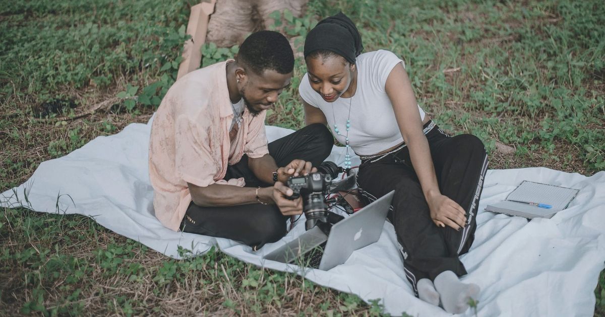 A couple sitting on a picnic blanket in a grassy area reviewing photos on a camera and laptop, enjoying leisure time outdoors.