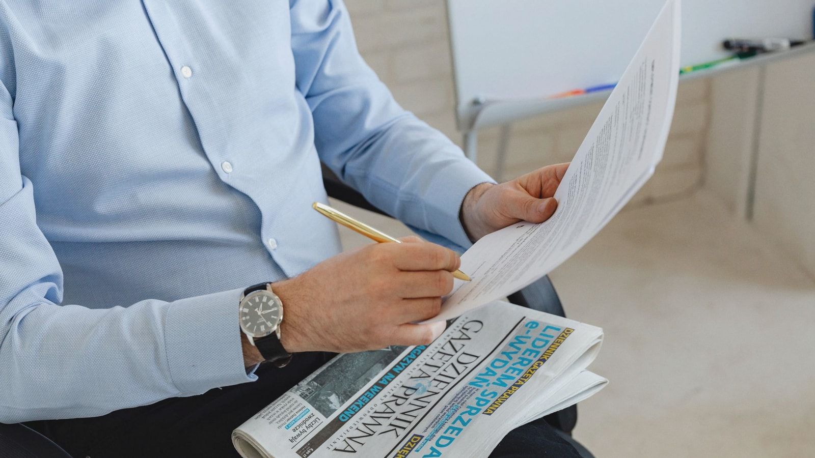 Businessman in blue shirt reviewing documents with a pen over a newspaper in an office setting.