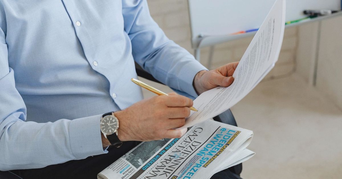 Businessman in blue shirt reviewing documents with a pen over a newspaper in an office setting.