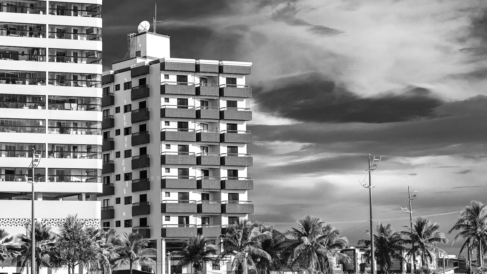 A couple walks along a sandy beach in front of apartment buildings and palm trees under a dramatic, cloudy sky.