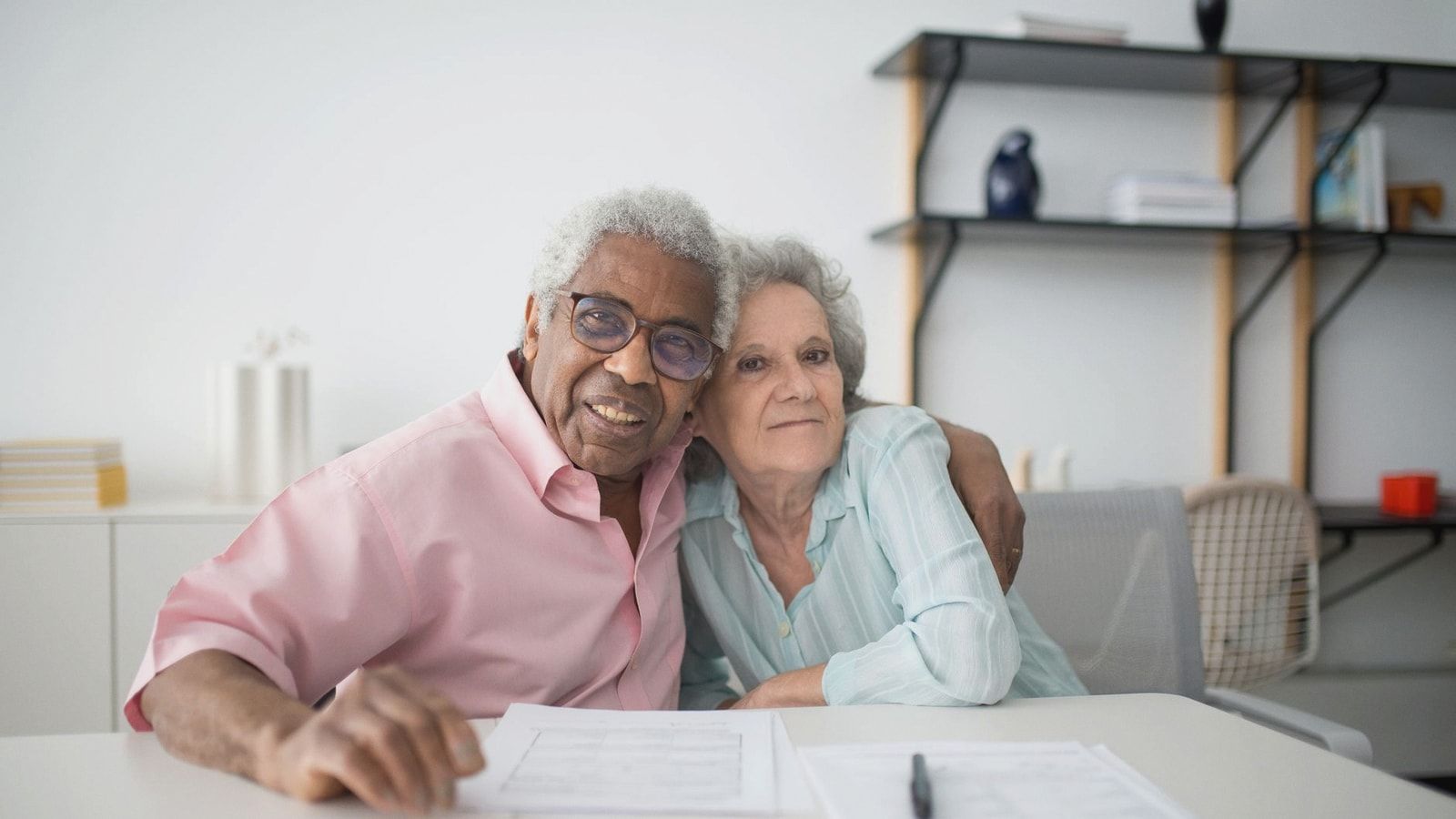 Elderly couple smiling and hugging while looking at documents on table indoors.