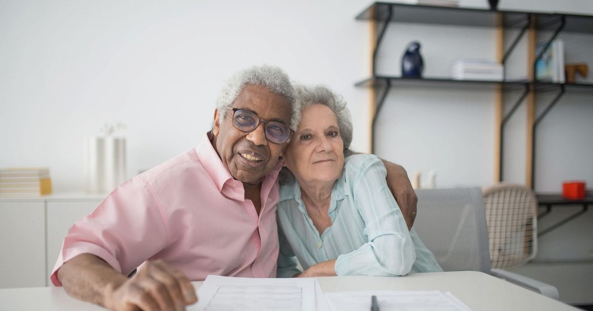 Elderly couple smiling and hugging while looking at documents on table indoors.