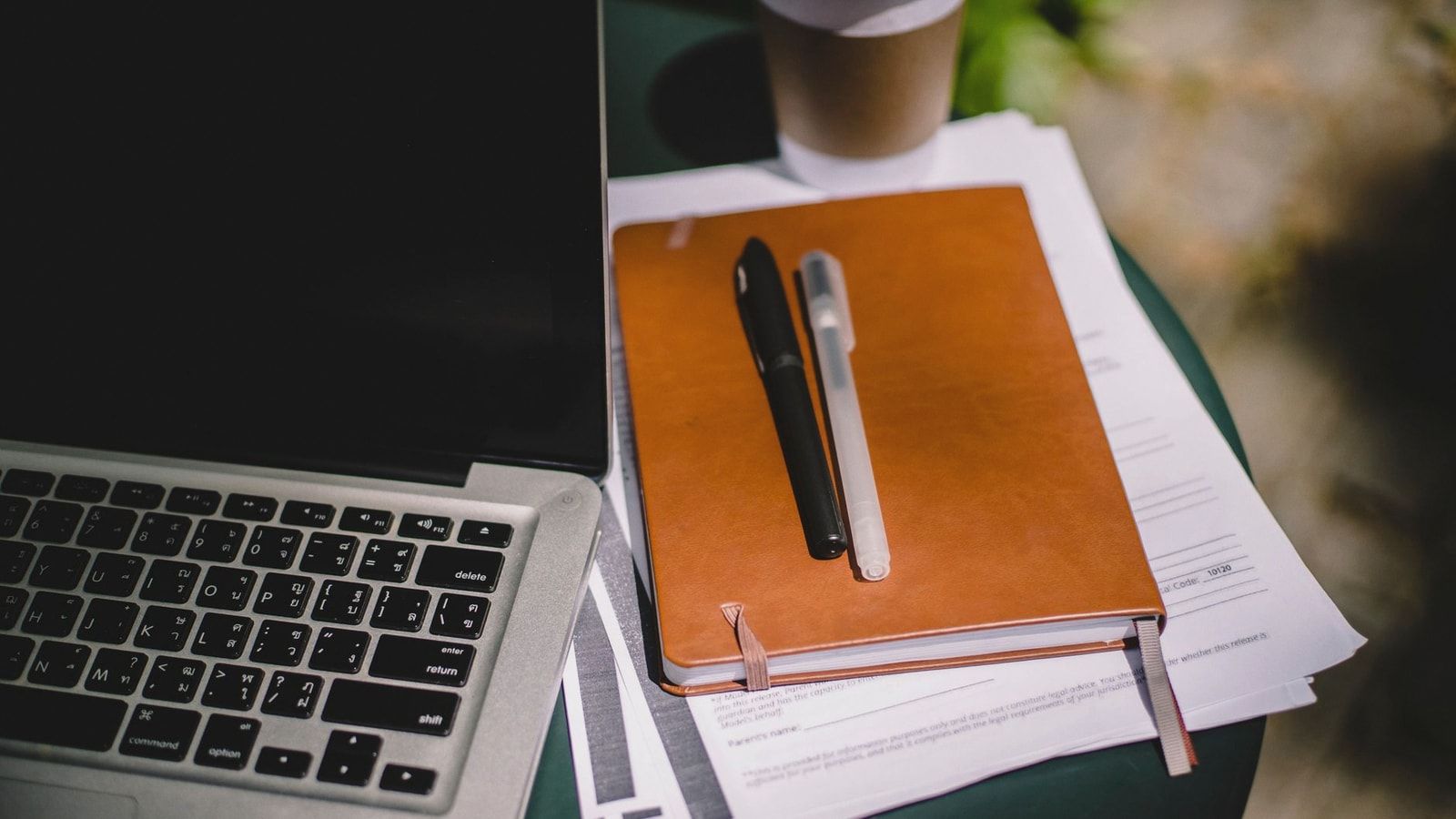 From above of leather notebook with papers and pens placed on table near laptop and cup of coffee