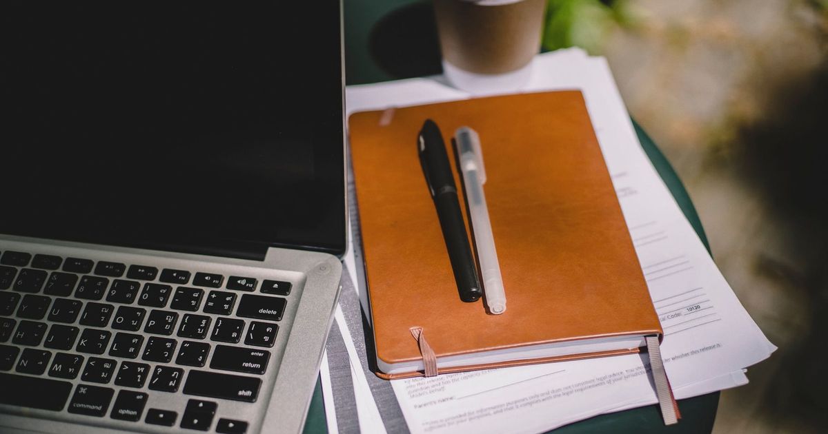 From above of leather notebook with papers and pens placed on table near laptop and cup of coffee