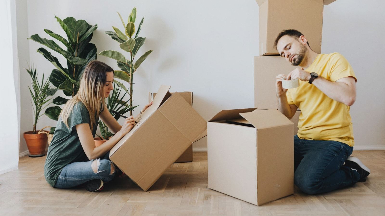 Side view full length glad couple packing stuff into cardboard boxes while sitting on floor at home during relocation in new house