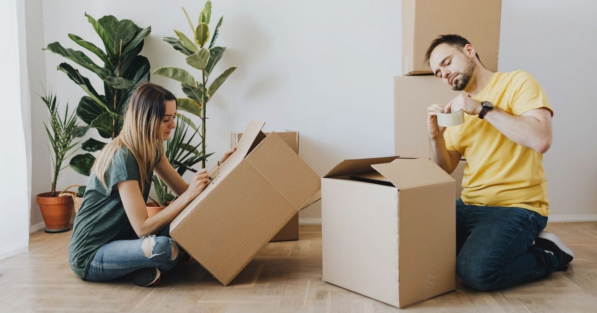 Side view full length glad couple packing stuff into cardboard boxes while sitting on floor at home during relocation in new house