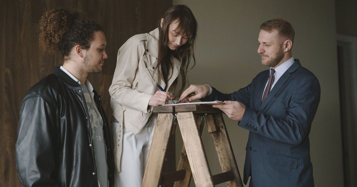 Three individuals signing a contract on a ladder inside a new house, symbolizing a real estate deal.