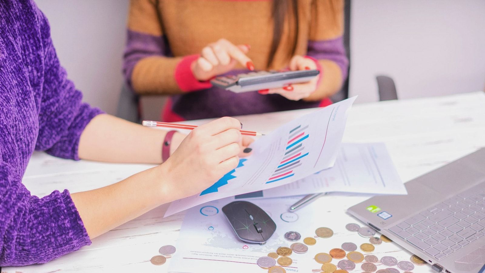 Two women analyzing financial charts with a laptop and coins on desk in an office setting.