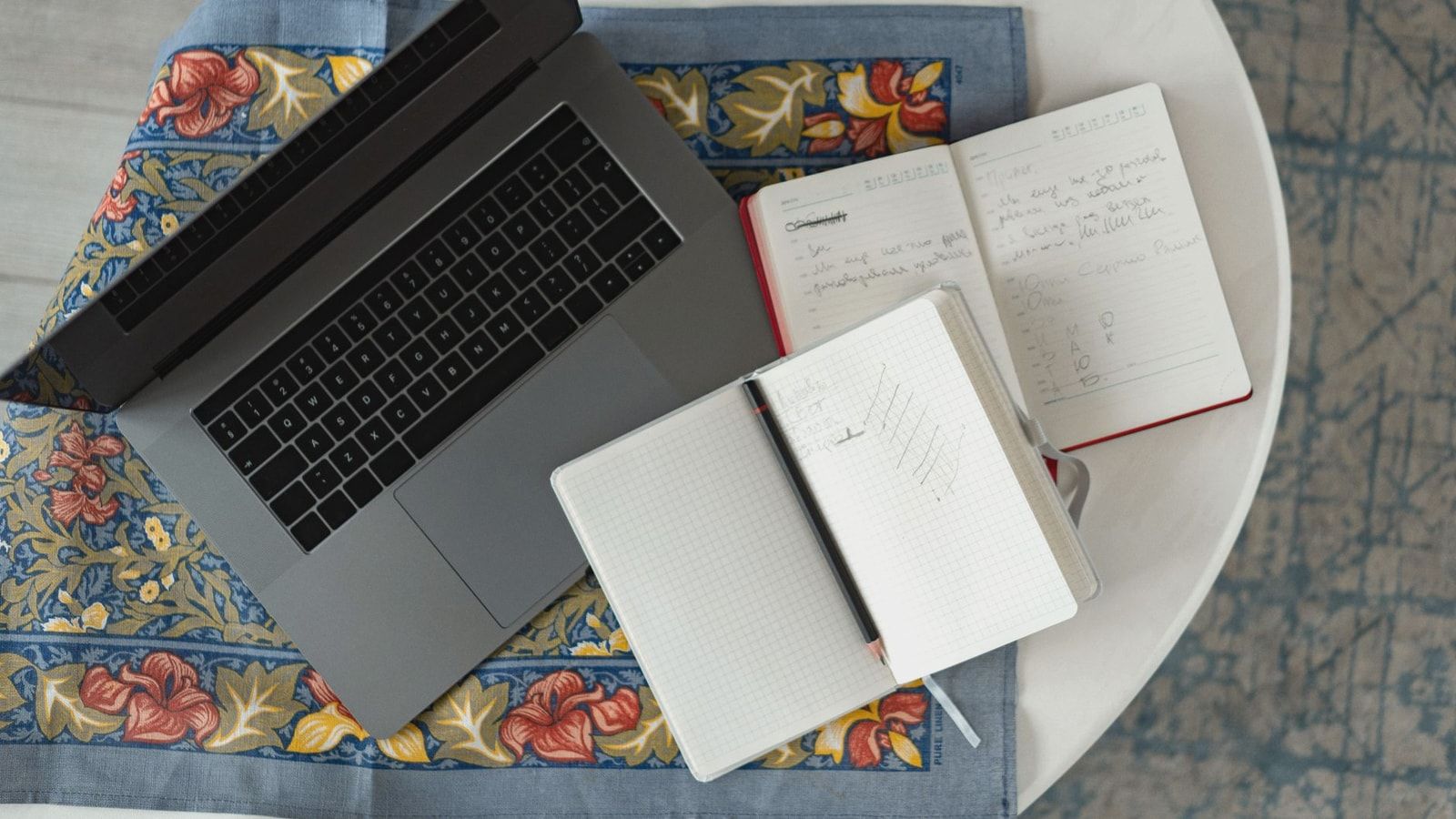 Overhead shot of a laptop with notebooks and a pencil on a patterned cloth.