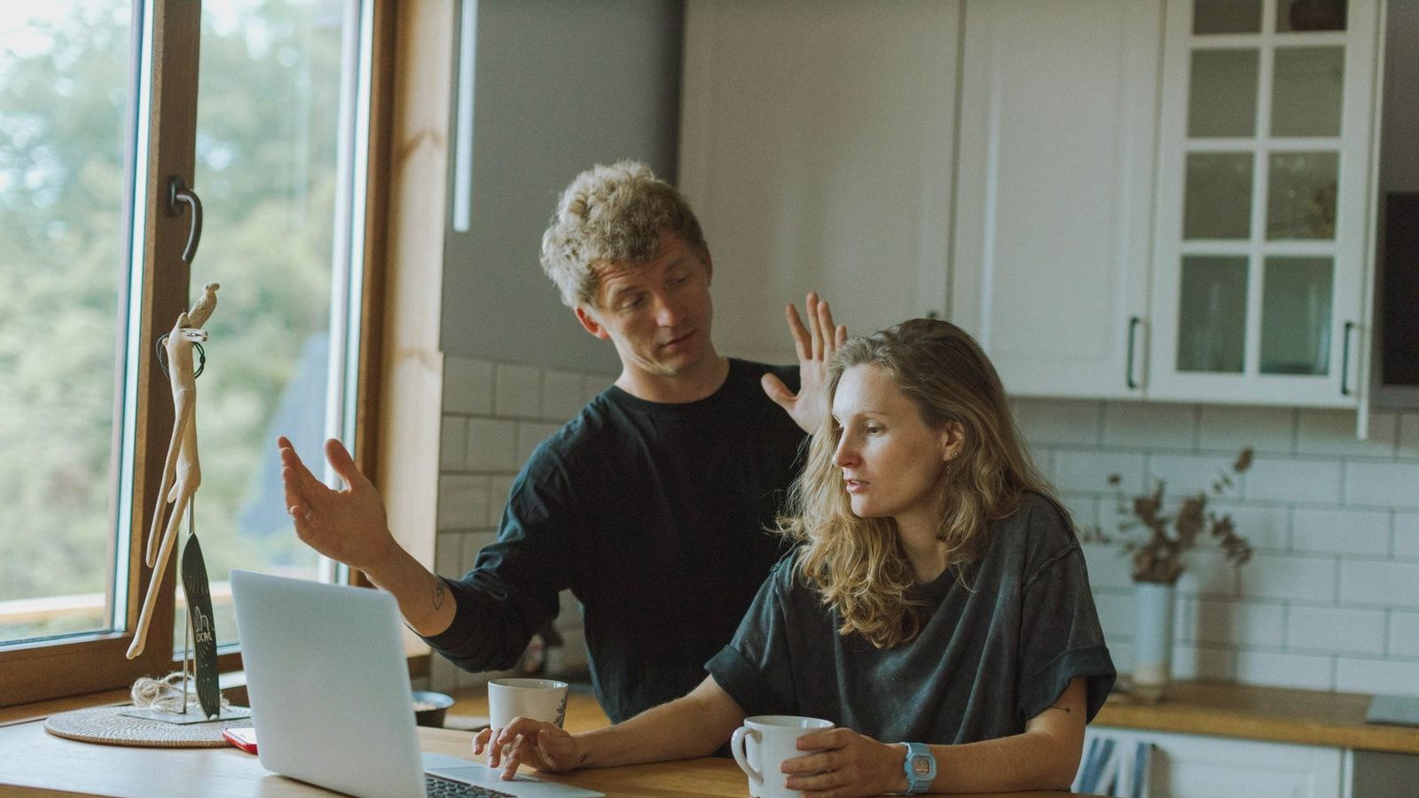 A couple interacting in a bright kitchen using a laptop at a wooden counter.