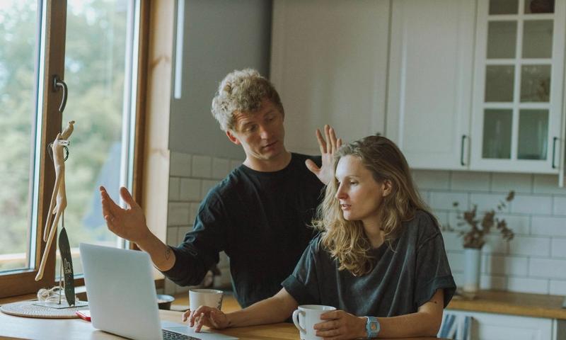 A couple interacting in a bright kitchen using a laptop at a wooden counter.