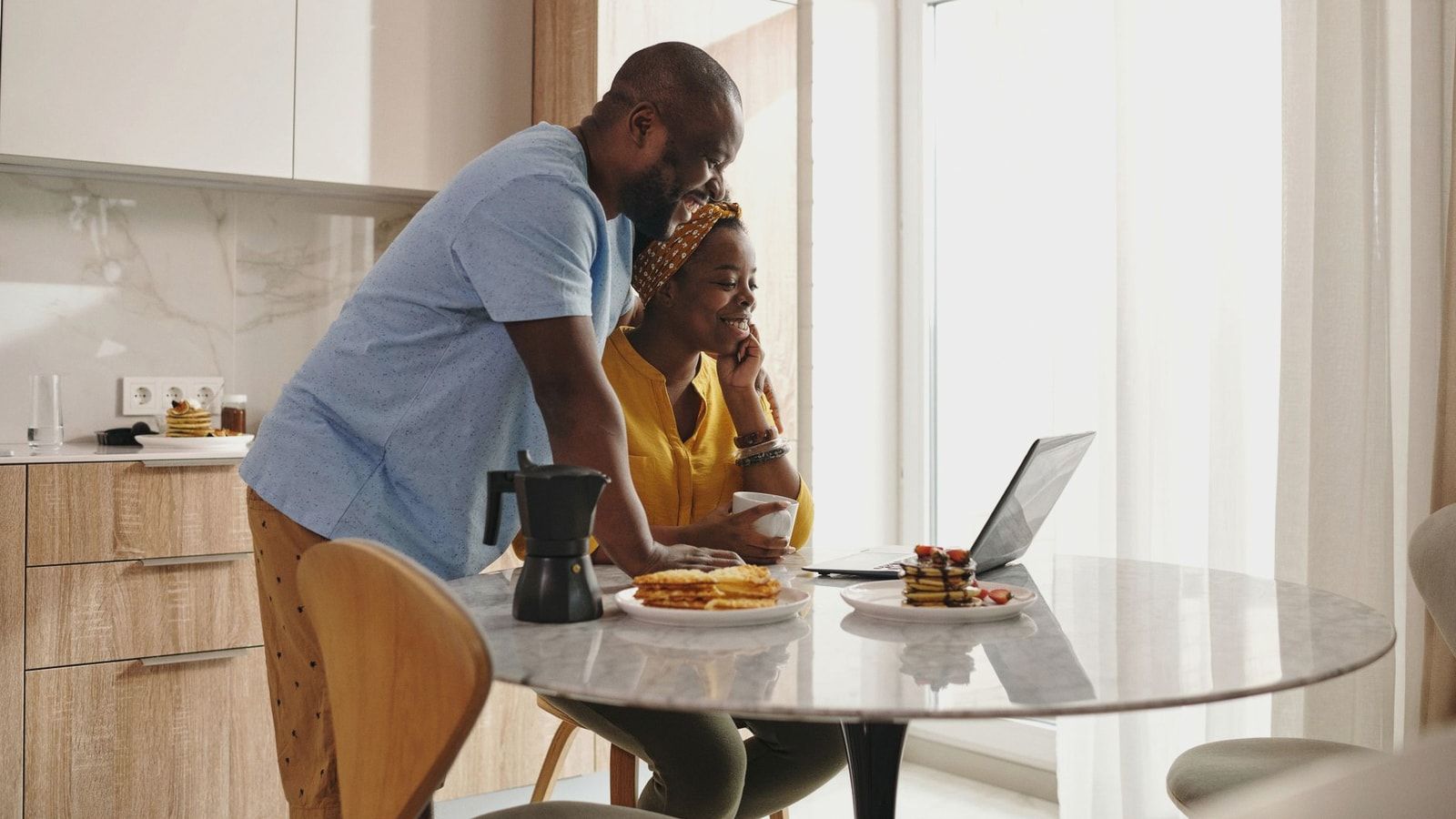 A joyful couple sharing breakfast and working on a laptop in their cozy kitchen.