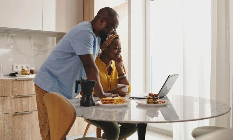 A joyful couple sharing breakfast and working on a laptop in their cozy kitchen.