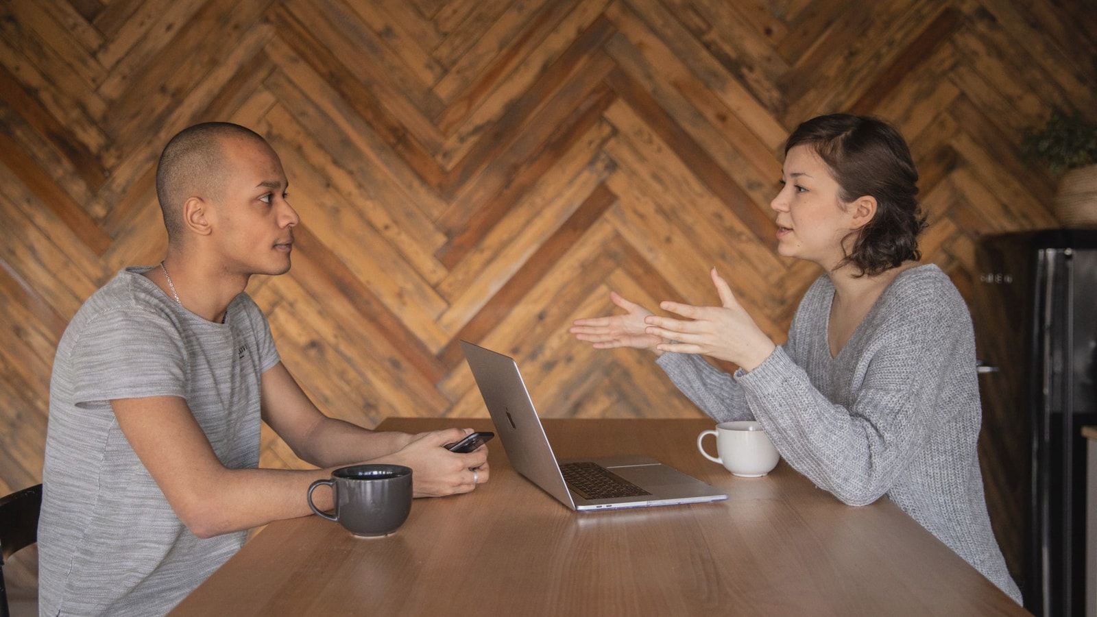 Focused woman explaining opinion to ethnic male coworker during business teamwork sitting at table with laptop and coffee cups in cozy kitchen against wooden wall and looking at each other