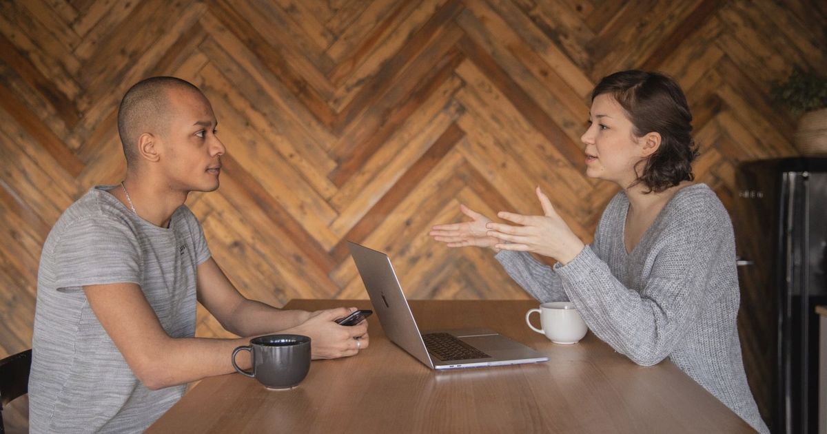 Focused woman explaining opinion to ethnic male coworker during business teamwork sitting at table with laptop and coffee cups in cozy kitchen against wooden wall and looking at each other