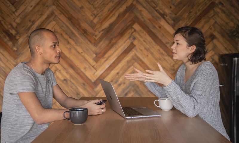 Focused woman explaining opinion to ethnic male coworker during business teamwork sitting at table with laptop and coffee cups in cozy kitchen against wooden wall and looking at each other