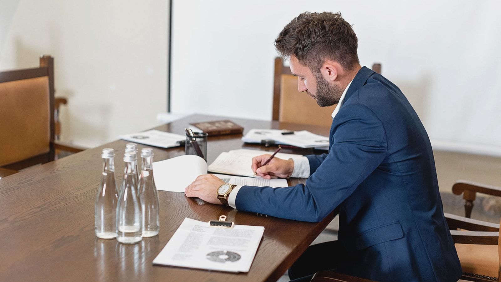 A businessman in a blue suit signing a contract at an office desk with paperwork and water bottles.