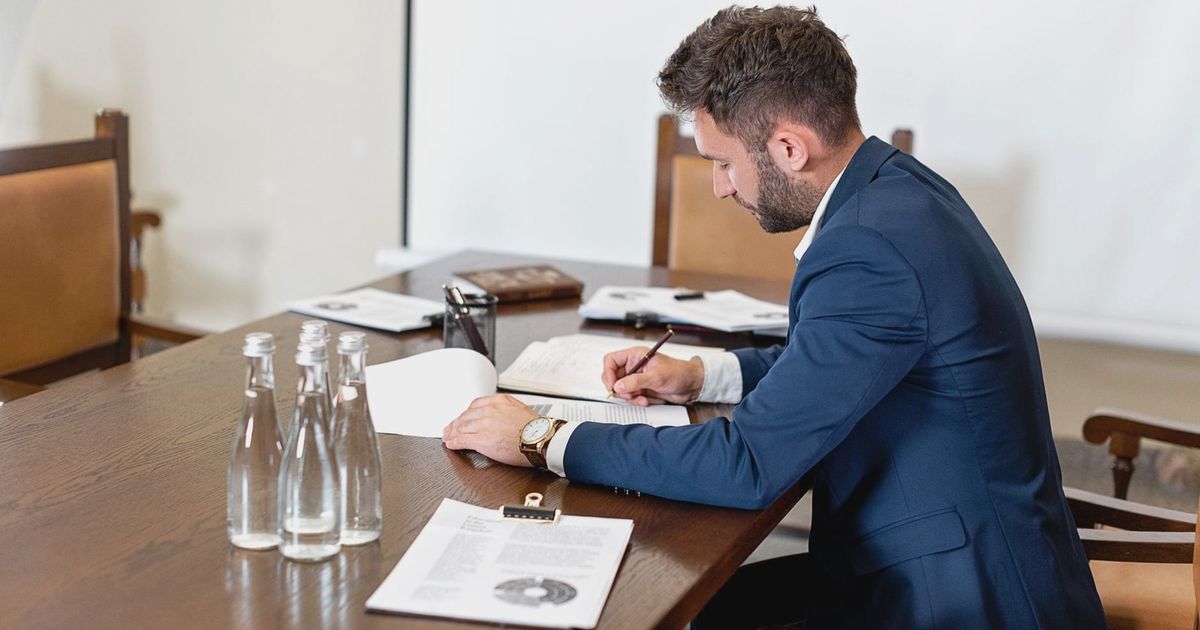 A businessman in a blue suit signing a contract at an office desk with paperwork and water bottles.