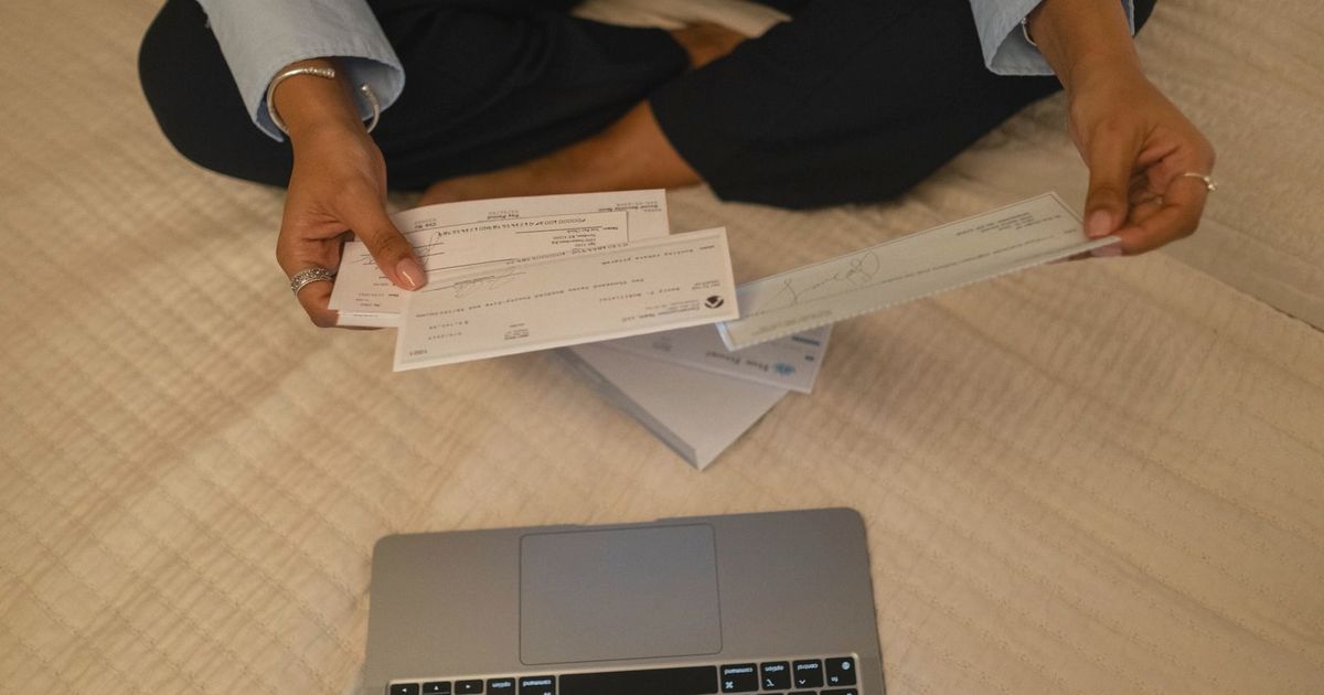 Close-up of hands holding cheques beside a laptop indoors for financial tasks.