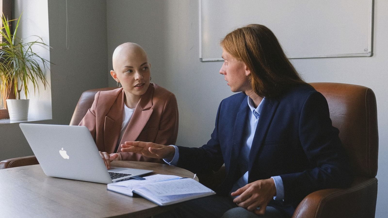 Confident young coworkers wearing formal jackets sitting in leather armchairs near table with netbook and organizer while sharing business ideas