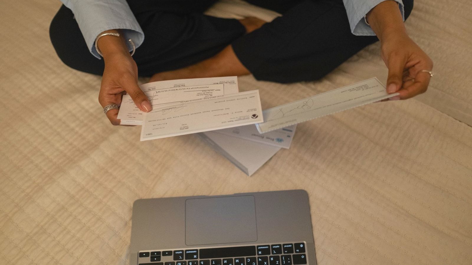Close-up of hands holding cheques beside a laptop indoors for financial tasks.