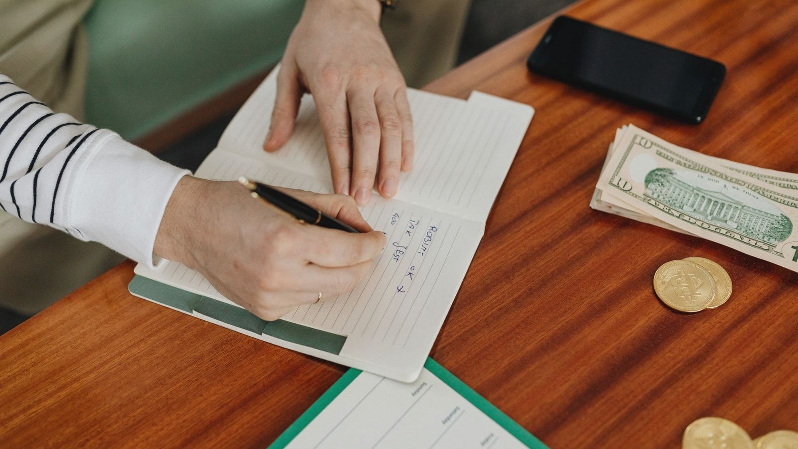 Person writing in a notebook on a wooden table with cash, coins, and a phone nearby.