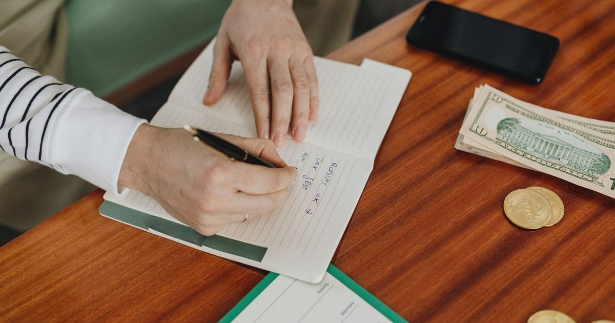 Person writing in a notebook on a wooden table with cash, coins, and a phone nearby.