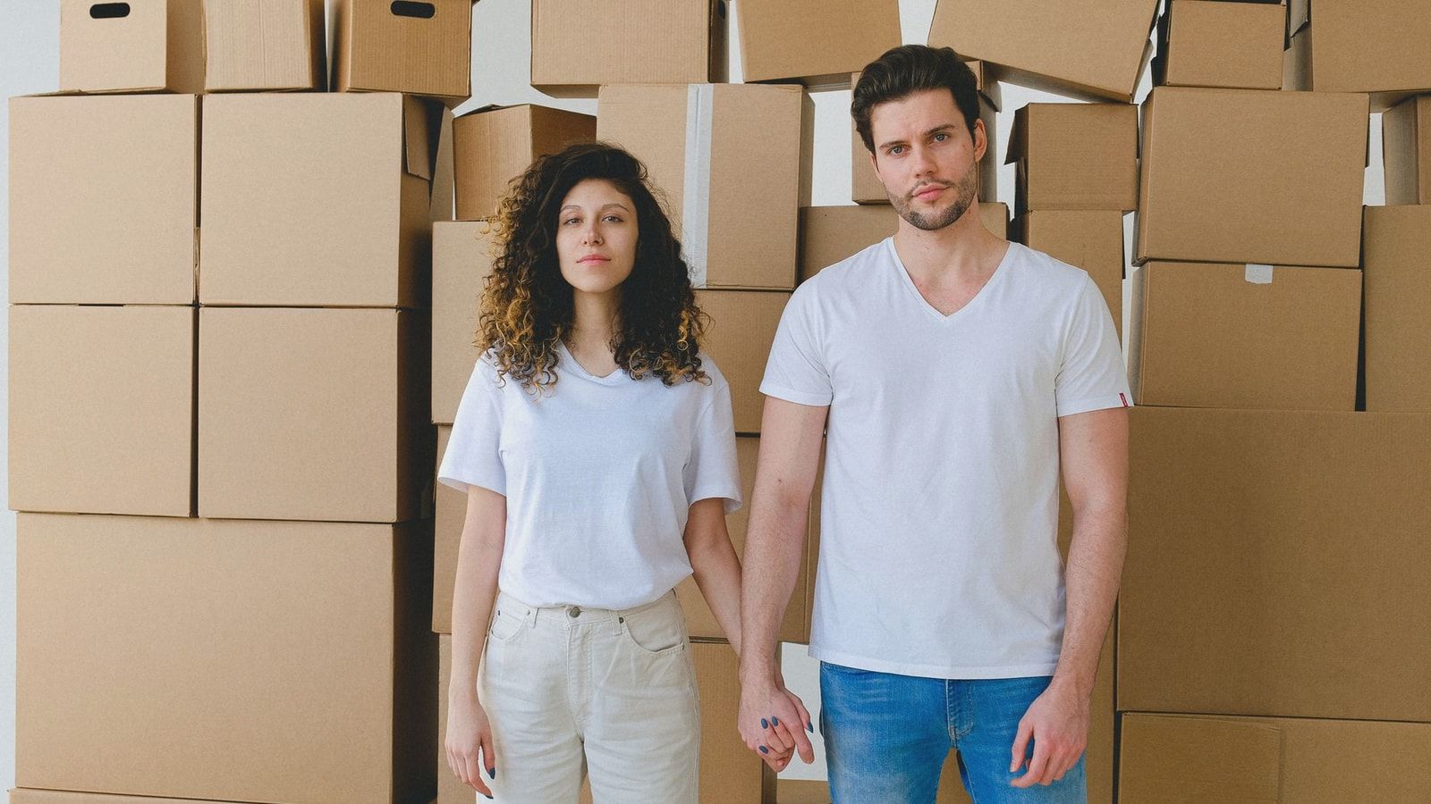 A young couple holding hands in front of a stack of cardboard boxes, possibly indicating moving or relocation.