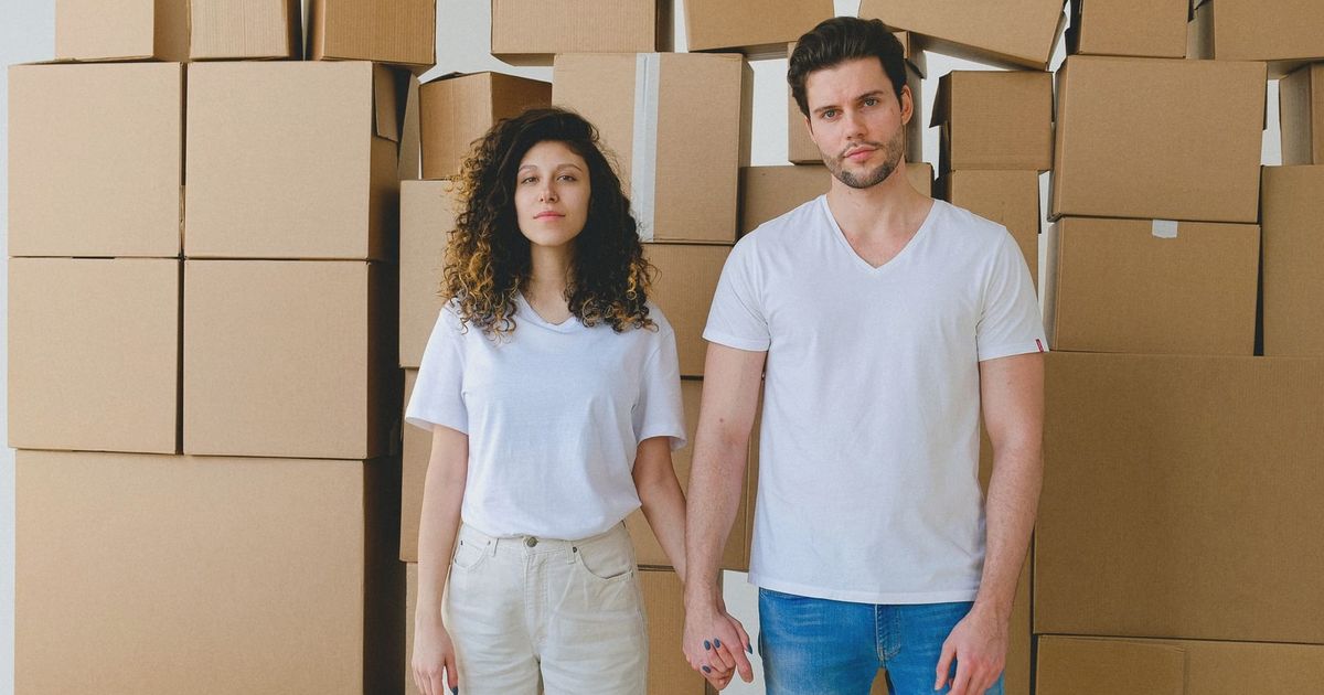 A young couple holding hands in front of a stack of cardboard boxes, possibly indicating moving or relocation.