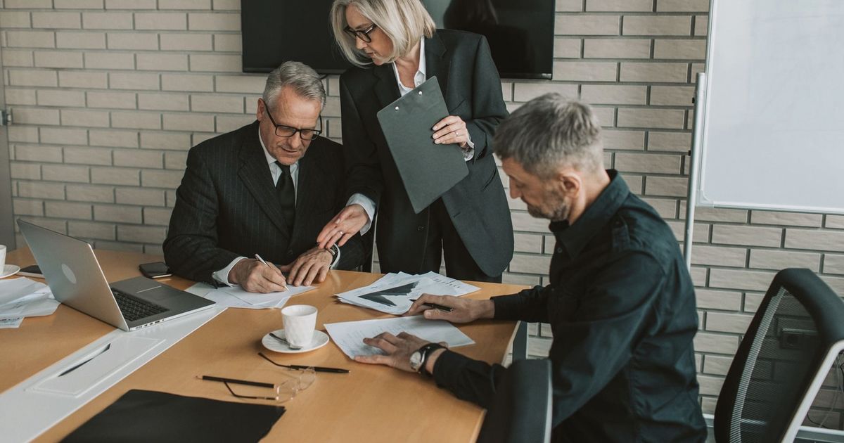 Three mature professionals in a business meeting discussing and signing documents in an office setting.