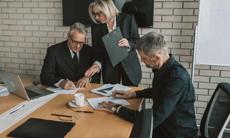 Three mature professionals in a business meeting discussing and signing documents in an office setting.