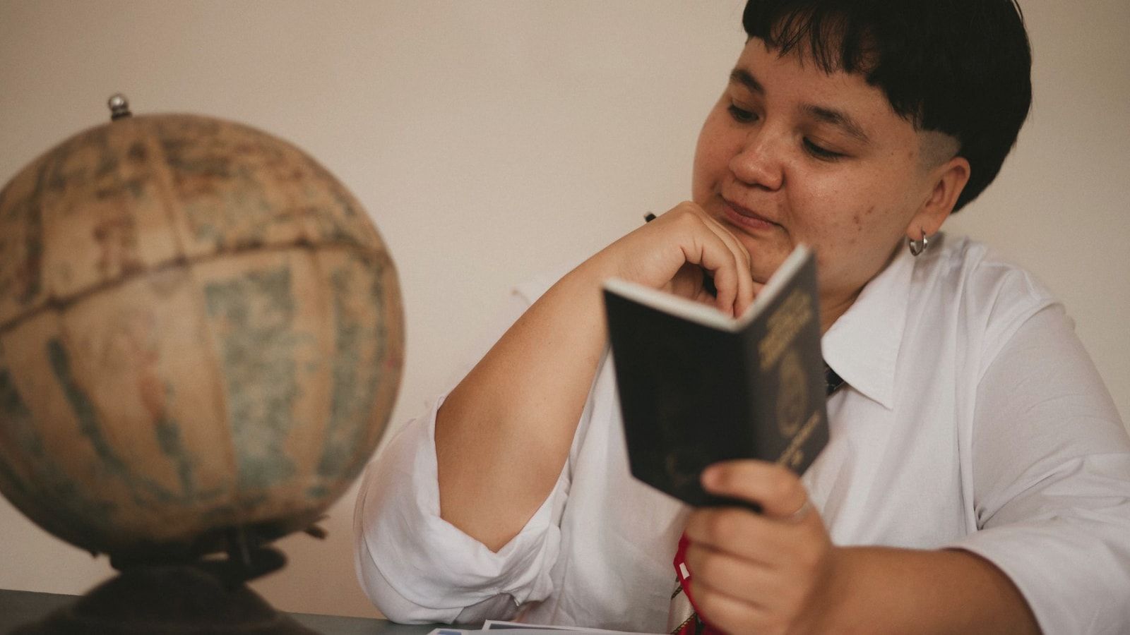 A pensive young adult reading a passport next to a vintage globe, symbolizing travel dreams.