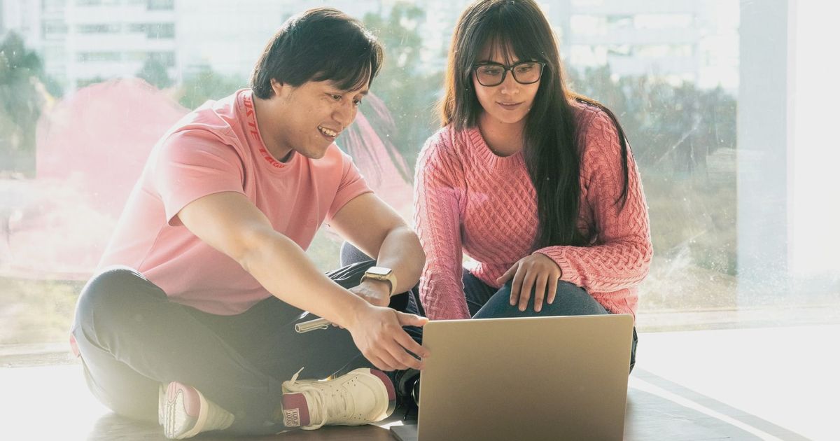 Two young adults in casual attire working together on a laptop by a window.