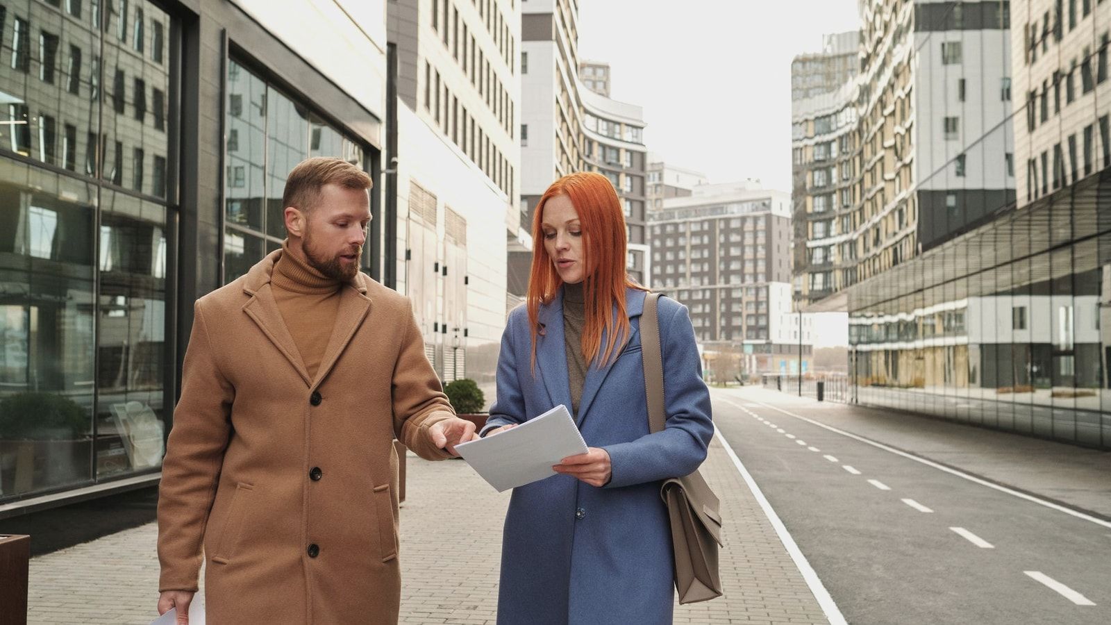 Two business professionals in winter attire reviewing documents in a modern cityscape.
