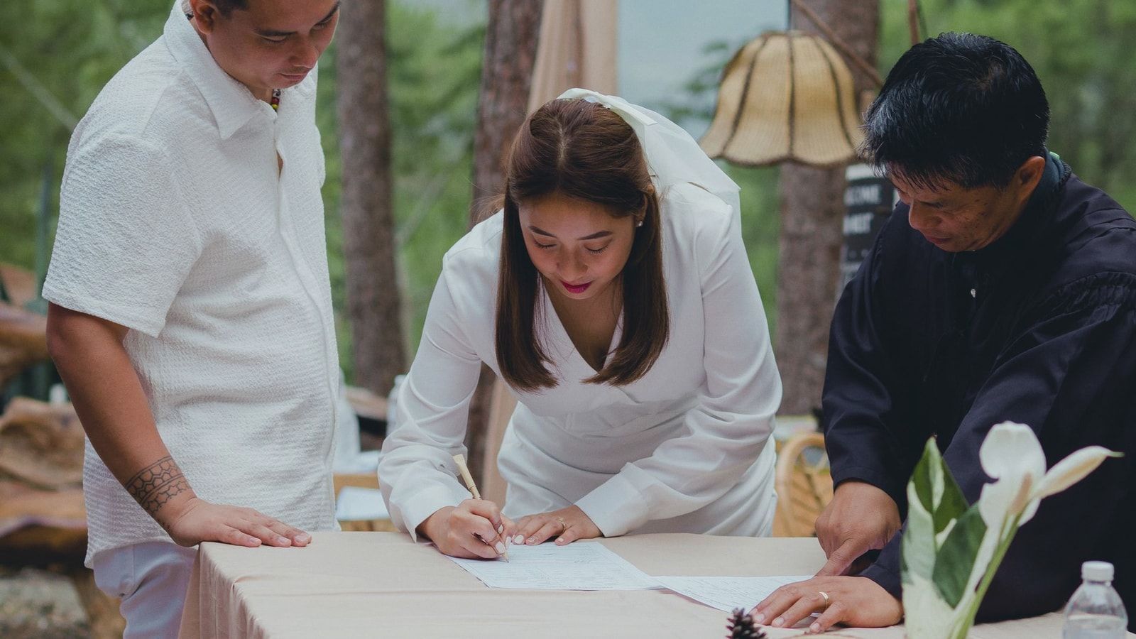 Bride signing marriage certificate surrounded by groom and officiant during outdoor wedding.