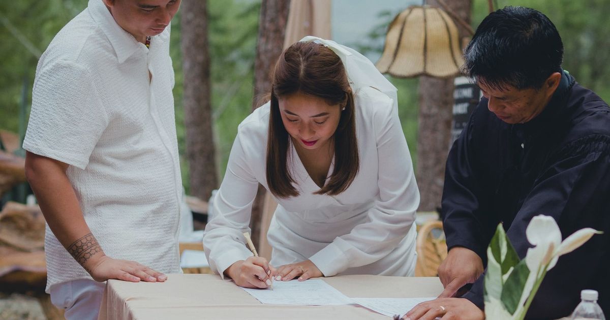 Bride signing marriage certificate surrounded by groom and officiant during outdoor wedding.