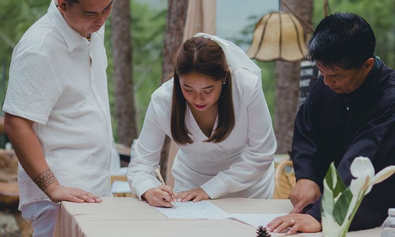 Bride signing marriage certificate surrounded by groom and officiant during outdoor wedding.