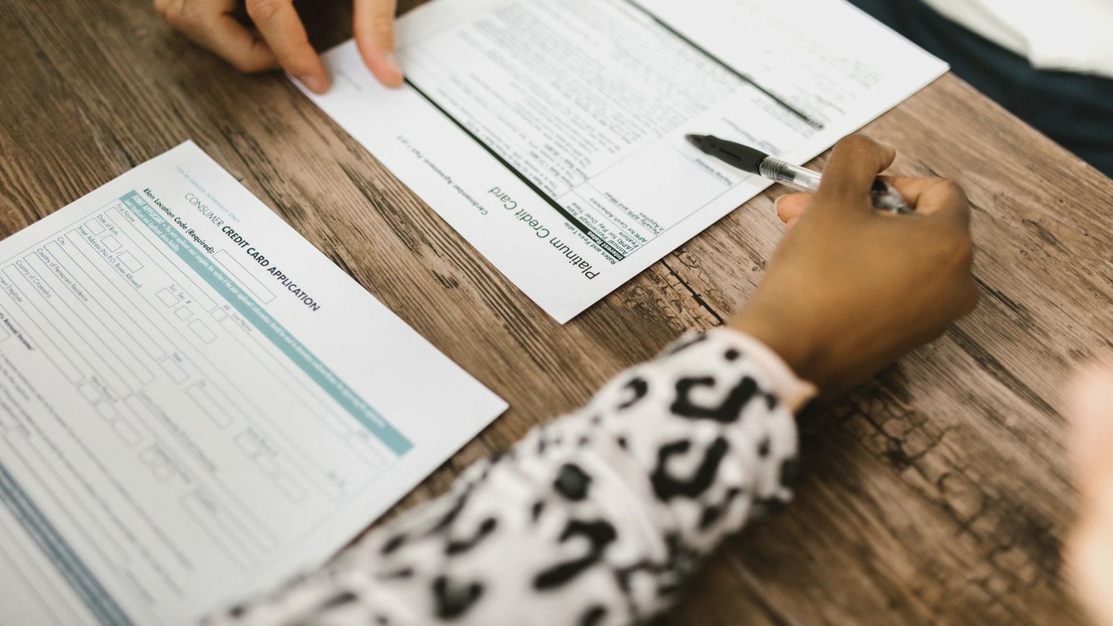 Close-up of two people reviewing and filling out a credit card application on a wooden table.