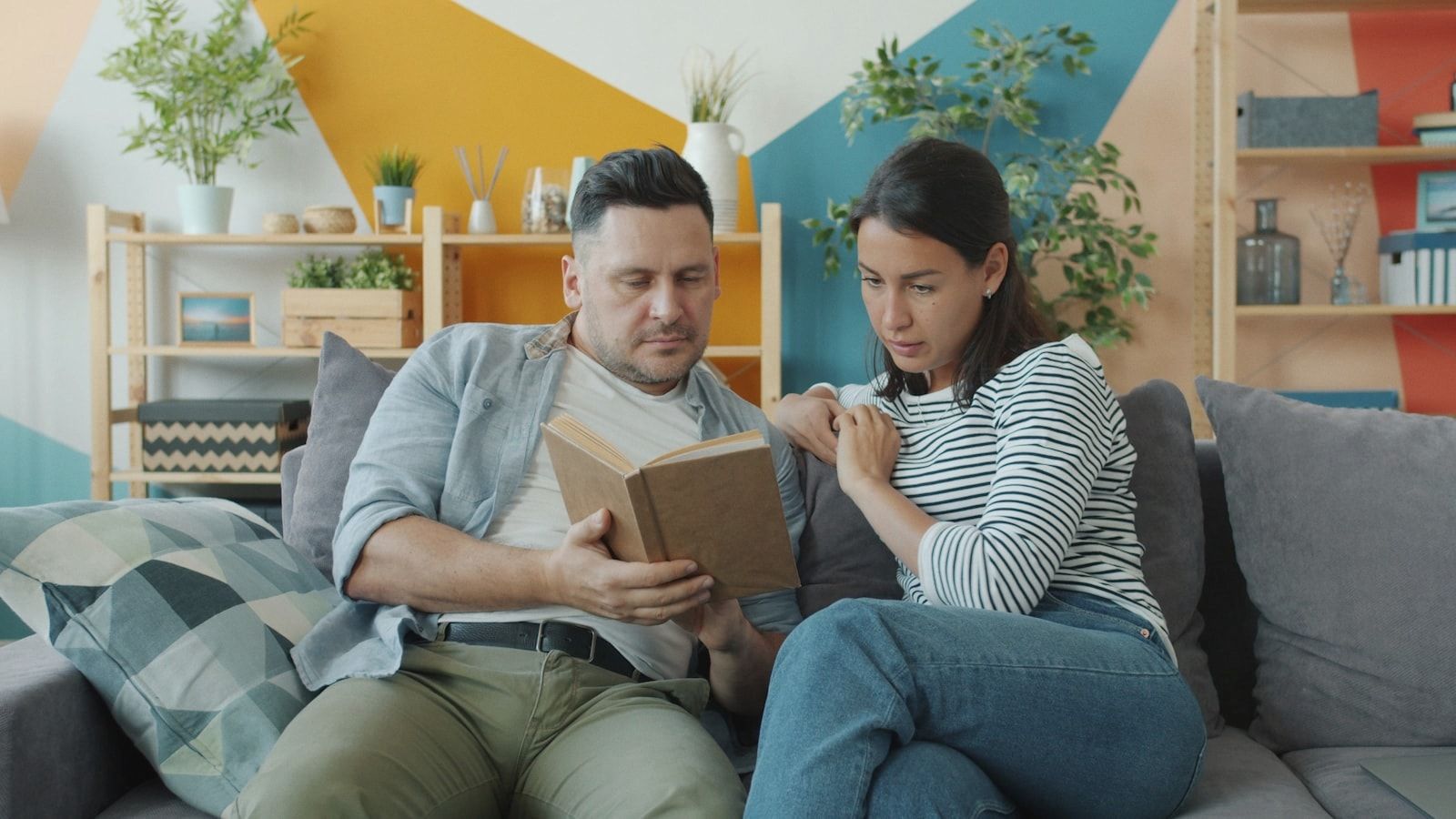 Young couple man and woman are reading book talking enjoying literature in apartment sitting on couch together. Youth culture and family relationship concept.