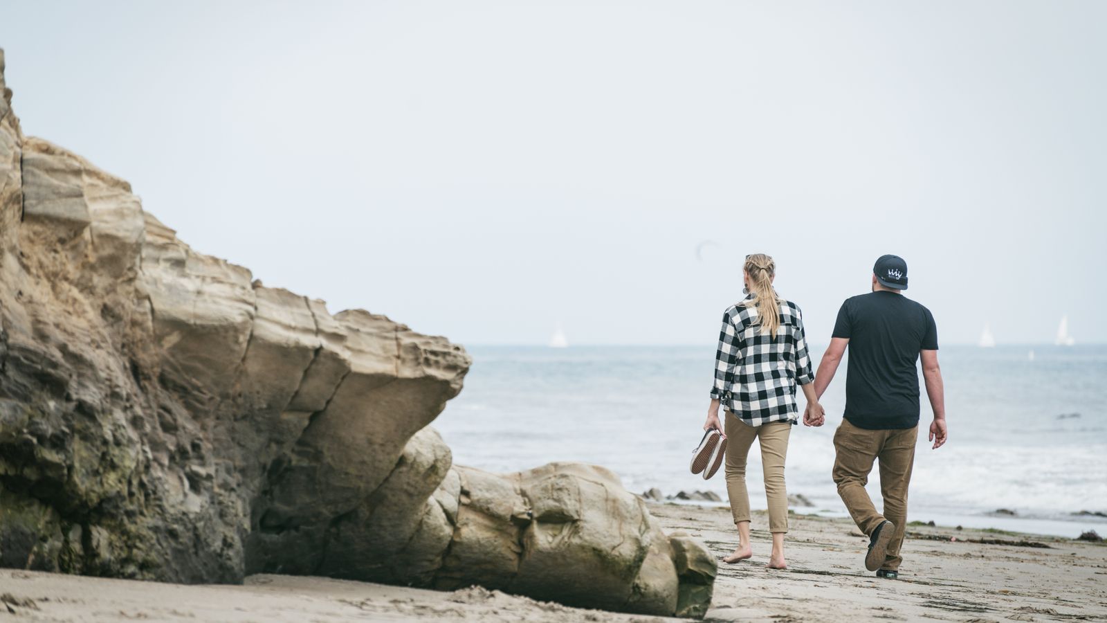 A couple holds hands walking along a California beach past coastal rocks, sailboats on the ocean behind them.